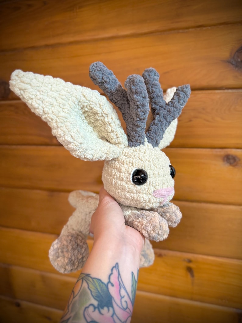 Pu&ograve; includere: Un peluche fatto a mano all'uncinetto a forma di creatura simile a un cervo. Il giocattolo presenta grandi orecchie flosce, corna grigie e occhi neri a bottone. Il corpo &egrave; color crema con accenti grigi e rosa.
