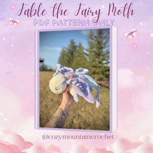 Op de afbeelding: Een gehaakte feeënmot in pastelkleuren blauw, paars en geel. De afbeelding toont de tekst "Fable the Fairy Moth PDF Pattern Only" en de Instagram-gebruikersnaam @cozymountaincrochet. De achtergrond toont bomen en een blauwe lucht.