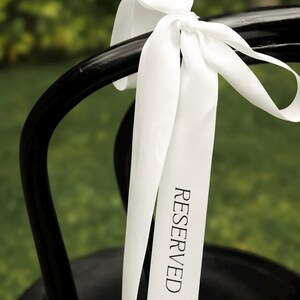 May include: A black chair with a white ribbon tied to the back. The ribbon has the word "RESERVED" printed on it in black. The background is a blurred green.