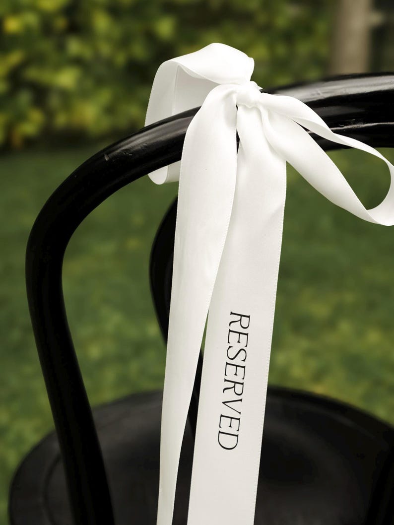 May include: A black chair with a white ribbon tied to the back. The ribbon has the word "RESERVED" printed on it in black. The background is a blurred green.
