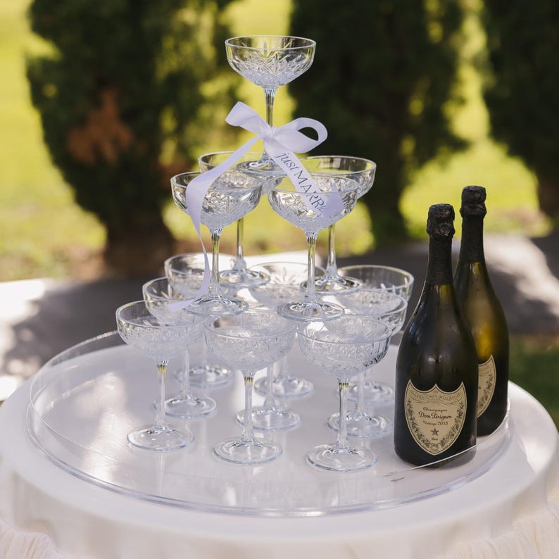 May include: A champagne tower made of crystal champagne flutes with a white ribbon that says "Just Married" and two bottles of Dom P&eacute;rignon champagne on a white round table.