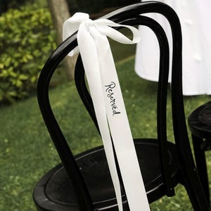 May include: Black wooden chair with a white ribbon tied to the back. The ribbon has the word "Reserved" printed on it in a script font. The chair is set on green grass, suggesting an outdoor event.