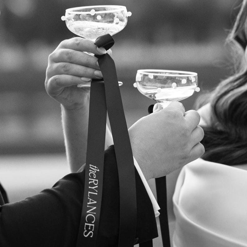 May include: Two clear glass champagne flutes with white polka dots, held by two hands. A black ribbon with the text "theRYLANCES" hangs from the flute in the left hand.