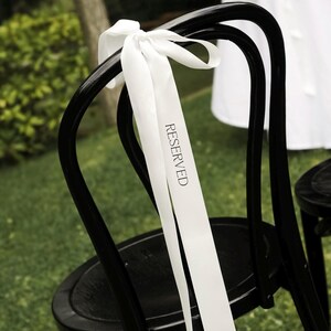 May include: Black wooden chair with a white ribbon tied to the back. The ribbon has the word "RESERVED" printed on it in black. The chair is on a green lawn, suggesting an outdoor event.