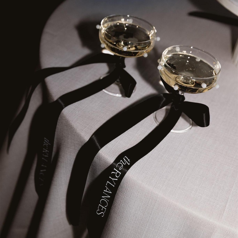May include: Two champagne flutes with pearl-like embellishments on the rims, tied with black satin ribbons that say "The Grylances" in white lettering.