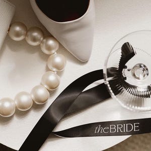 May include: A white shoe, a pearl necklace, black ribbon with the text "theBRIDE" printed on it, and a clear glass goblet.