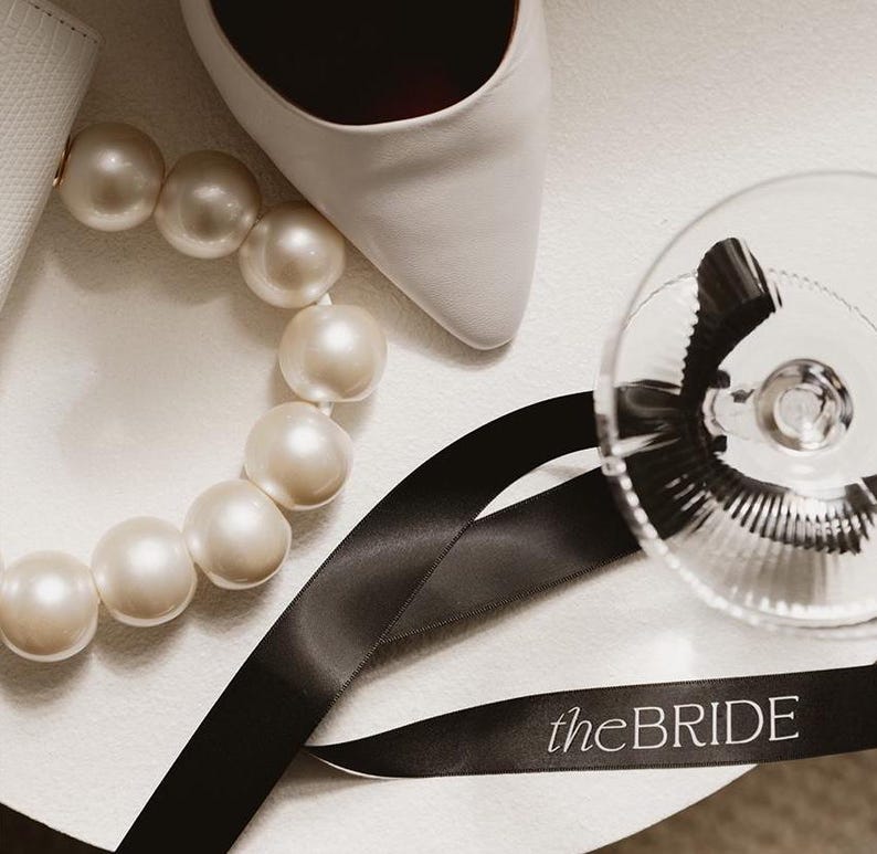 May include: A white shoe, a pearl necklace, black ribbon with the text "theBRIDE" printed on it, and a clear glass goblet.