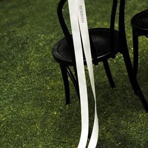 May include: Black wooden chair with a white ribbon tied to the back. The ribbon has the word "RESERVED" printed on it. The chair is on a green grassy surface.