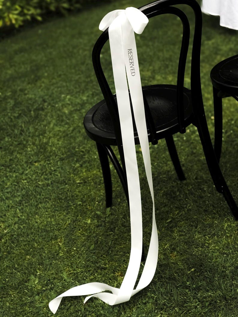 May include: Black wooden chair with a white ribbon tied to the back. The ribbon has the word "RESERVED" printed on it. The chair is on a green grassy surface.