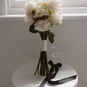 May include: A white rose bouquet tied with a white ribbon, sitting on a white table with a black ribbon that says "the bride".