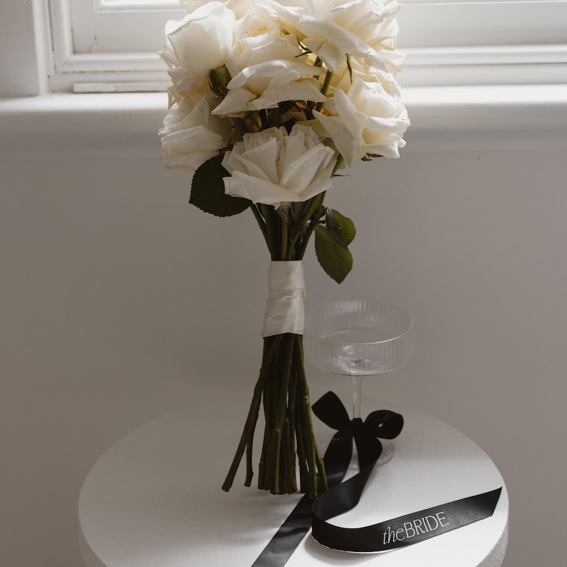 May include: A white rose bouquet tied with a white ribbon, sitting on a white table with a black ribbon that says "the bride".