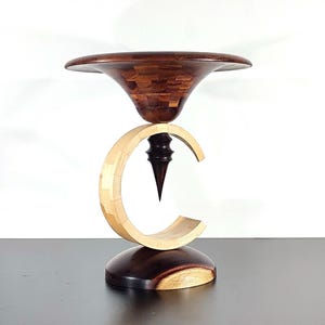 Puede incluir: Una escultura de madera hecha a mano con una parte superior marrón oscuro tipo mosaico, un centro en forma de media luna de color claro y una base marrón oscuro. La escultura tiene un diseño artístico único y se apoya sobre una superficie oscura.