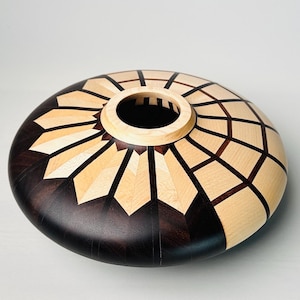 May include: A handcrafted wooden bowl with a dark brown base and a light wood and dark wood geometric pattern on the top. The bowl has a circular opening in the center.