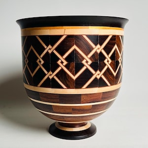 Puede incluir: Un jarrón de madera hecho a mano con borde y base marrón oscuro. El cuerpo presenta un patrón geométrico de formas entrelazadas en tonos de madera claros y oscuros. El jarrón mide aproximadamente 25 cm de alto.