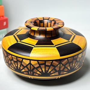 Puede incluir: Un jarrón decorativo de madera con un patrón geométrico. El jarrón presenta una combinación de madera clara y oscura, creando un contraste visual llamativo. El diseño incluye una base circular y una abertura acampanada, mostrando la artesanía.
