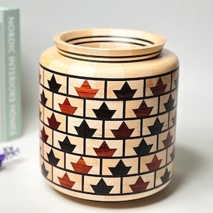 Può includere: Un vaso decorativo in legno di forma cilindrica. Il vaso presenta un motivo ripetuto di forme a forma di foglia nere e rosse su uno sfondo di legno chiaro. Il vaso è alto circa 20 cm.