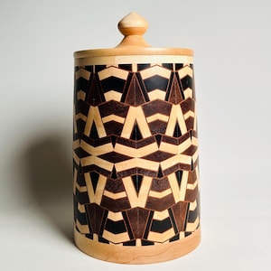 Può includere: Un contenitore cilindrico in legno con coperchio. Il corpo presenta un motivo geometrico nelle tonalità del marrone, nero e legno naturale. Il coperchio e la base sono di una tonalità di legno più chiara e il pomello ha una forma arrotondata.