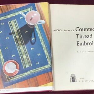 May include: An open book featuring a blue embroidered tray cloth and a pink teapot and teacup. The book's cover reads "Anchor Book of Counted Thread Embroidery". The tray cloth has a geometric design with white and yellow accents.