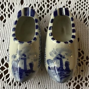 May include: Two ceramic clogs, white with blue windmill and floral patterns. The rims are decorated with blue stripes and dots. These decorative clogs are a charming addition to any home decor.