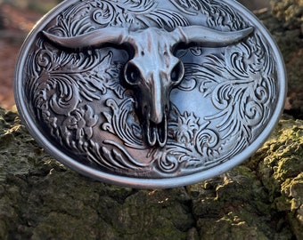 May include: Oval silver belt buckle with a steer skull design. The buckle has detailed floral and scrollwork patterns. The background shows a blurred view of a tree and sky. The buckle is made of metal.