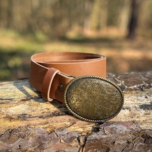 May include: A tan leather belt featuring a large, oval-shaped brass buckle. The buckle has a detailed, embossed floral pattern across its surface and a twisted rope-style border. The belt strap is smooth, light brown leather with a single loop.