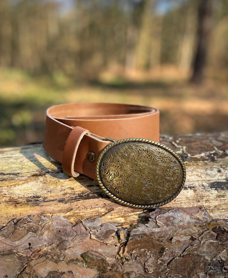 May include: A tan leather belt featuring a large, oval-shaped brass buckle. The buckle has a detailed, embossed floral pattern across its surface and a twisted rope-style border. The belt strap is smooth, light brown leather with a single loop.