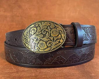 Peut inclure: Une ceinture en cuir marron avec une boucle ornée dorée à motif floral. La ceinture est embossée avec un motif de montagne et d'oiseau. Le texte "ELLIOT HOLDEN Leather" est visible en bas de l'image.