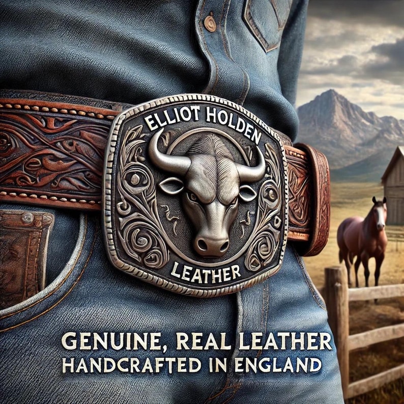May include: A silver belt buckle with a bull's head design and the text "Elliot Holden Leather" on a brown leather belt. The belt is worn by a person wearing blue jeans. The background shows a mountain range and a wooden fence.