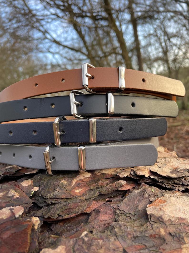 May include: Four leather belts in different colors, stacked on top of each other. The belts are brown, black, dark blue, and gray. Each belt has a silver buckle.