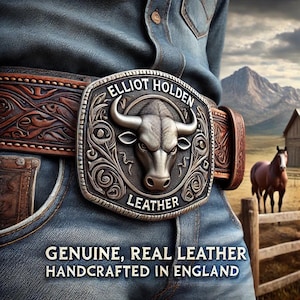 May include: A brown leather belt with a large silver buckle featuring a bull's head design. The buckle is engraved with the words "ELLIOT HOLDEN LEATHER." The belt is worn with blue denim jeans. The text "GENUINE, REAL LEATHER HANDCRAFTED IN ENGLAND" is visible.