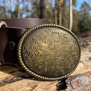 May include: A dark brown leather belt featuring an oval-shaped brass buckle. The buckle has a rope-textured border and is deeply engraved with a detailed floral pattern across its entire surface. A single metal rivet secures the leather strap.