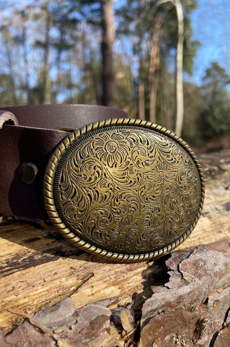 May include: A dark brown leather belt featuring an oval-shaped brass buckle. The buckle has a rope-textured border and is deeply engraved with a detailed floral pattern across its entire surface. A single metal rivet secures the leather strap.