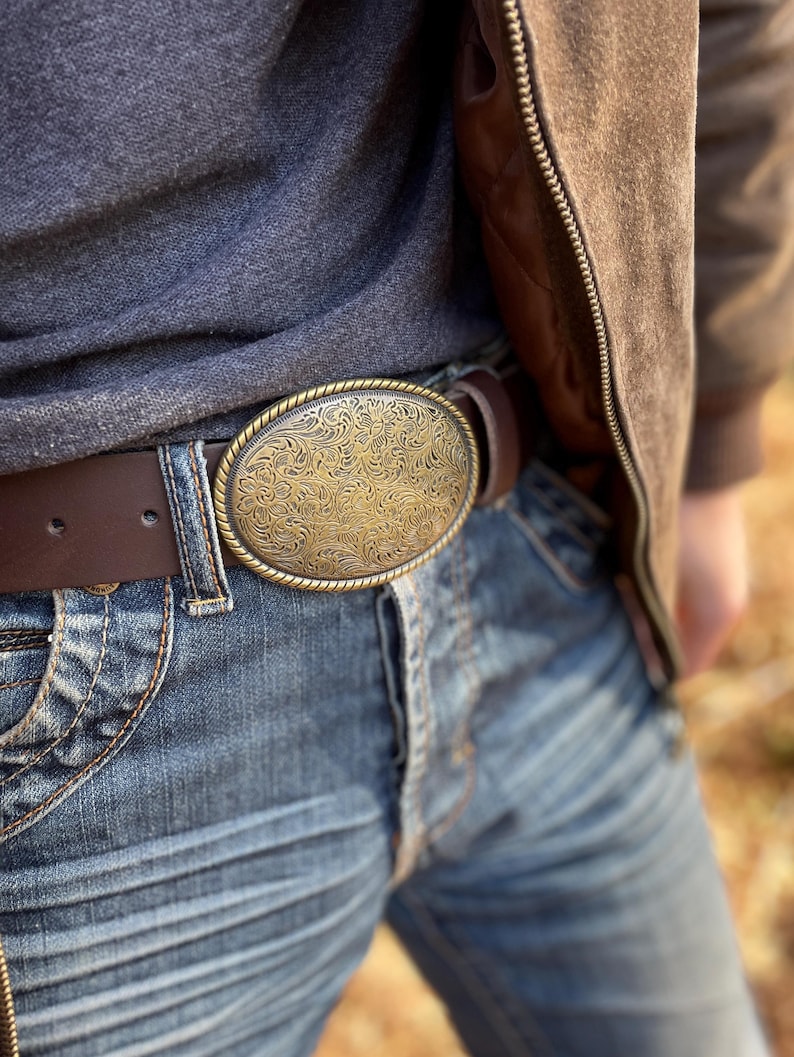 May include: An oval-shaped brass-toned belt buckle featuring an intricate, embossed floral pattern and a twisted rope-style border. The buckle is attached to a dark brown leather belt worn with blue denim jeans and a dark gray shirt.