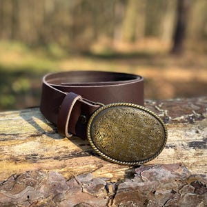 May include: A dark brown leather belt featuring an oval-shaped brass buckle. The buckle has a rope-style border and is decorated with an intricate, embossed floral pattern across its entire surface. The belt includes a matching leather keeper loop.
