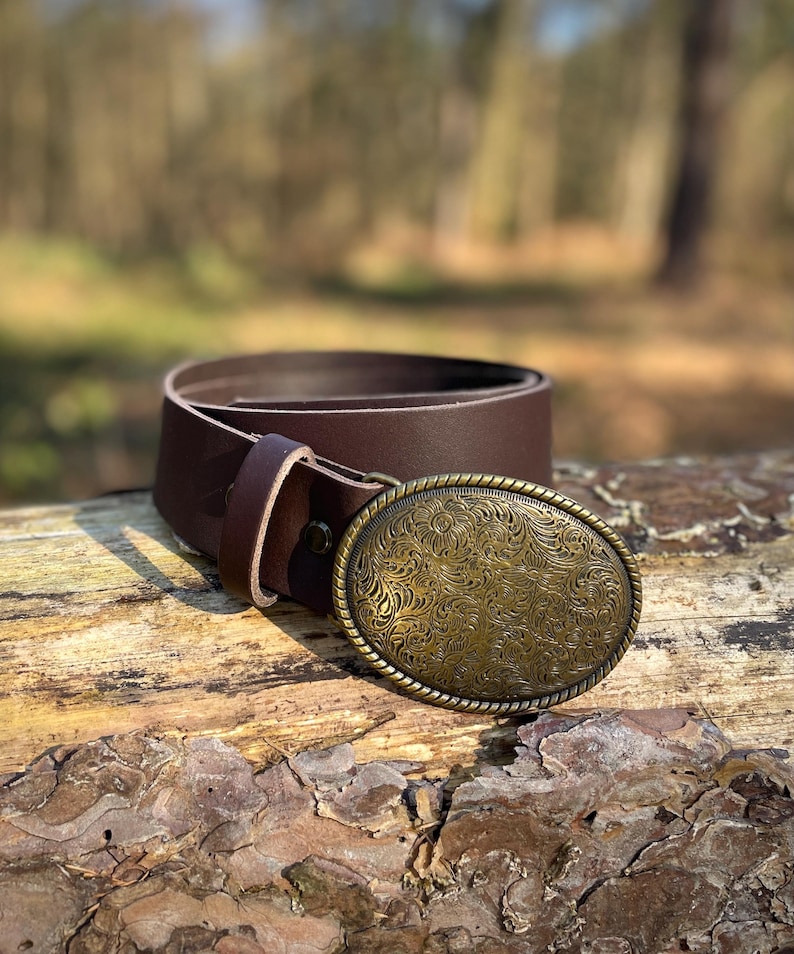 May include: A dark brown leather belt featuring an oval-shaped brass buckle. The buckle has a rope-style border and is decorated with an intricate, embossed floral pattern across its entire surface. The belt includes a matching leather keeper loop.