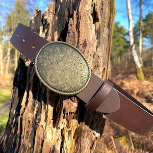May include: A dark brown leather belt featuring a large, oval-shaped brass buckle. The buckle has a rope-style border and is decorated with an intricate, embossed floral pattern across its entire surface. The belt includes a matching leather loop.