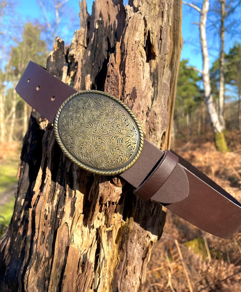 May include: A dark brown leather belt featuring a large, oval-shaped brass buckle. The buckle has a rope-style border and is decorated with an intricate, embossed floral pattern across its entire surface. The belt includes a matching leather loop.