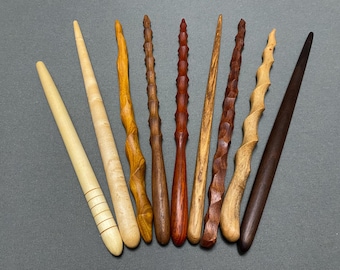 May include: A collection of eight handcrafted wooden wands in various shades of brown and tan. The wands vary in design, with some featuring spiral, ridged, and smooth textures. The wands are arranged on a gray surface.