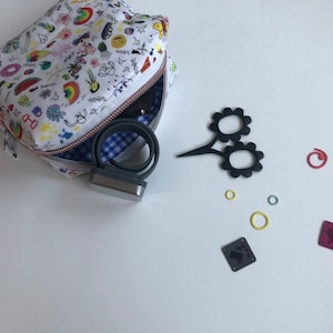 May include: A white cosmetic bag with a colorful doodle pattern, open to reveal a gray device and a blue and white gingham lining. Black flower-shaped scissors and various small, colorful rings and squares are scattered nearby.