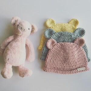 May include: A plush pink pig toy stands next to three crocheted hats with bear ears. The hats are in pastel colors: yellow, blue-green, and pink. The hats are handmade and have a tag on the brim.