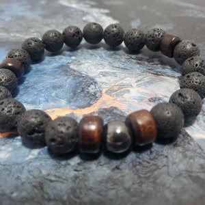 May include: A beaded bracelet featuring black lava rock beads, interspersed with brown wooden beads and a single silver-colored bead. The bracelet is strung on an elastic band, showcasing a natural, earthy aesthetic.