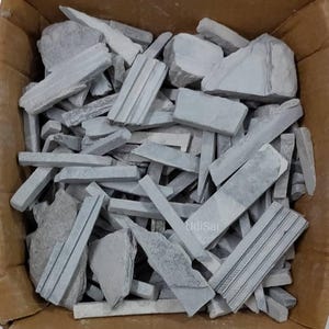 Op de afbeelding: Een kartonnen doos gevuld met grijze leistenen potloden en stukken. De leistenen stukken variëren in grootte en vorm, sommige tonen gegraveerde lijnen. De afbeelding is van bovenaf genomen, waarbij de textuur en kleur van de leisteen worden getoond.
