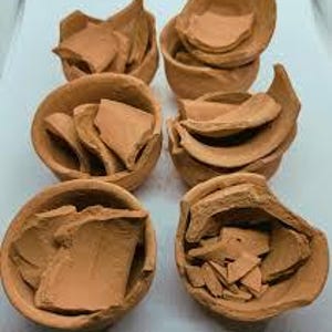 Op de afbeelding: Zes kleine, ongeglazuurde terracotta kommen, elk gevuld met gebroken stukken van hetzelfde materiaal. De kommen zijn gerangschikt in twee rijen van drie, met verschillende vormen en maten van de gebroken fragmenten.