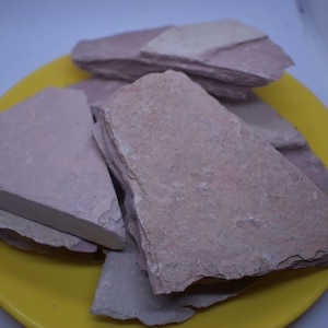 Pode incluir: Uma coleção de pedaços de pedra de cor rosa empoeirado de forma irregular é disposta em um prato amarelo brilhante. As pedras têm uma superfície áspera e texturizada e tamanhos variados, sugerindo uma origem natural. O prato oferece um fundo contrastante.