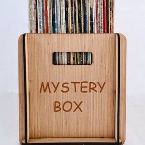 May include: A wooden box with a black trim and the words "MYSTERY BOX" printed on the front. The box is filled with a variety of vintage vinyl records.