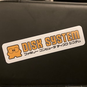 May include: A black zippered case with a white sticker that says "DISK SYSTEM" and "ファミリーコンピュータディスクシステム" in Japanese.