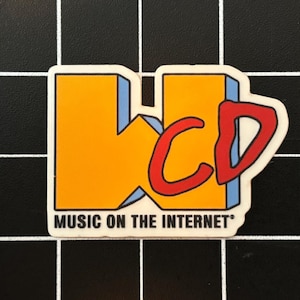 Może przedstawiać: Naklejka w kolorze żółtym i niebieskim z tekstem "WCD Music on the Internet" w stylizowanym kroju pisma. Litera "C" w "WCD" jest czerwona i częściowo zasłonięta przez "W".