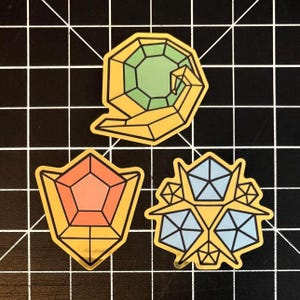 May include: Three geometric stickers with gold borders. One sticker features a green gem, another an orange gem, and the third has blue gems. All stickers have black outlines and are on a black grid background.