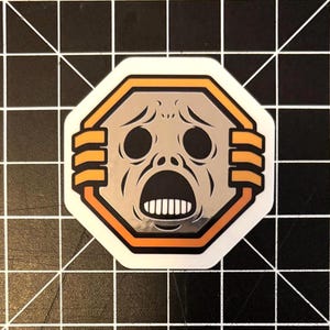May include: A sticker featuring a distressed face with wide eyes and an open mouth, set within a silver and orange octagonal frame. The design has a cartoonish, slightly eerie aesthetic, with a white border.