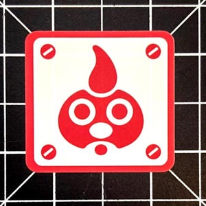 May include: A square sticker with a red border and a white center. The center features a red cartoon-like character with a flame-shaped head, two large eyes, and a small mouth. Four red rivets are in each corner.